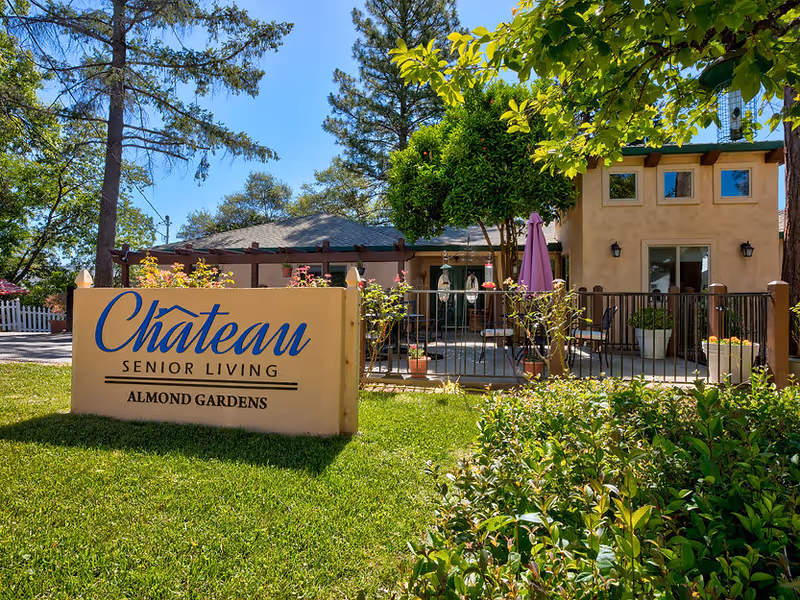Exterior view of Chateau Senior Living: Almond Gardens facility showing a beige sign with blue and black text in front of a green lawn and garden. Behind the sign is a fenced patio area with outdoor furniture, potted plants, and a purple umbrella. Trees and a clear blue sky are visible in the background.