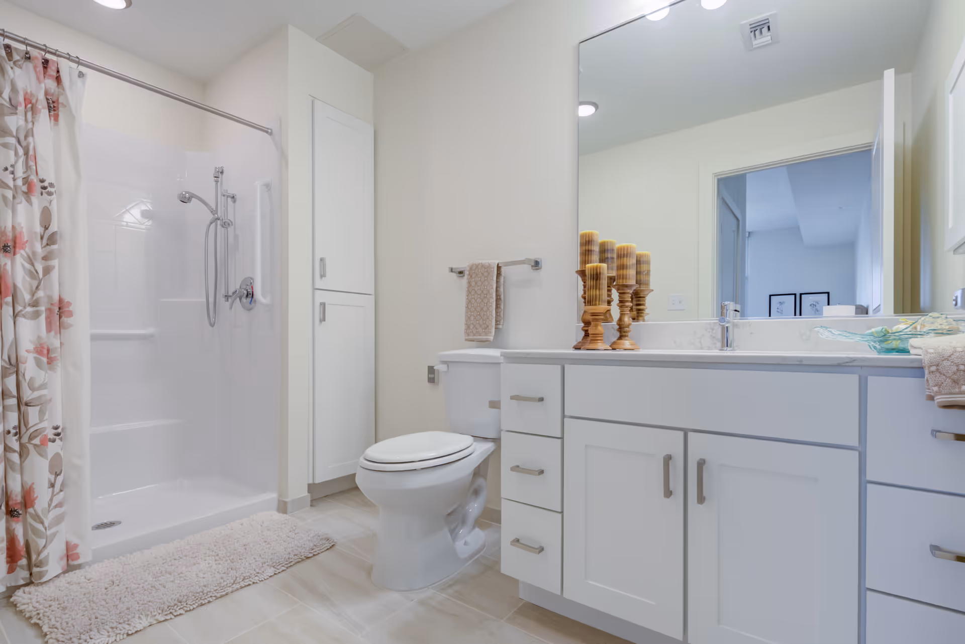 A clean and modern bathroom featuring a white toilet, a white vanity with drawers and cabinets, a large mirror above the sink, and a shower with a floral-patterned curtain. The floor is tiled, and there is a beige bath mat in front of the shower. Decorative candles are placed on the vanity countertop.
