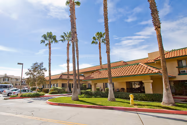 Exterior view of a single-story assisted living facility with a red tile roof, beige walls, and several tall palm trees in front. There is a driveway and parking area with cars visible on the left side under a partly cloudy blue sky.