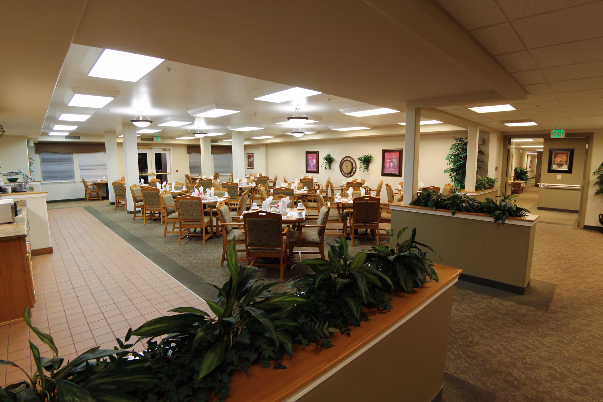 A spacious dining room in a senior living facility with multiple tables and chairs arranged neatly. Each table is set with white napkins and tableware. The room is well-lit with ceiling lights and has plants along a half wall in the foreground. There are framed pictures and wall decorations on the walls, and a hallway is visible to the right.
