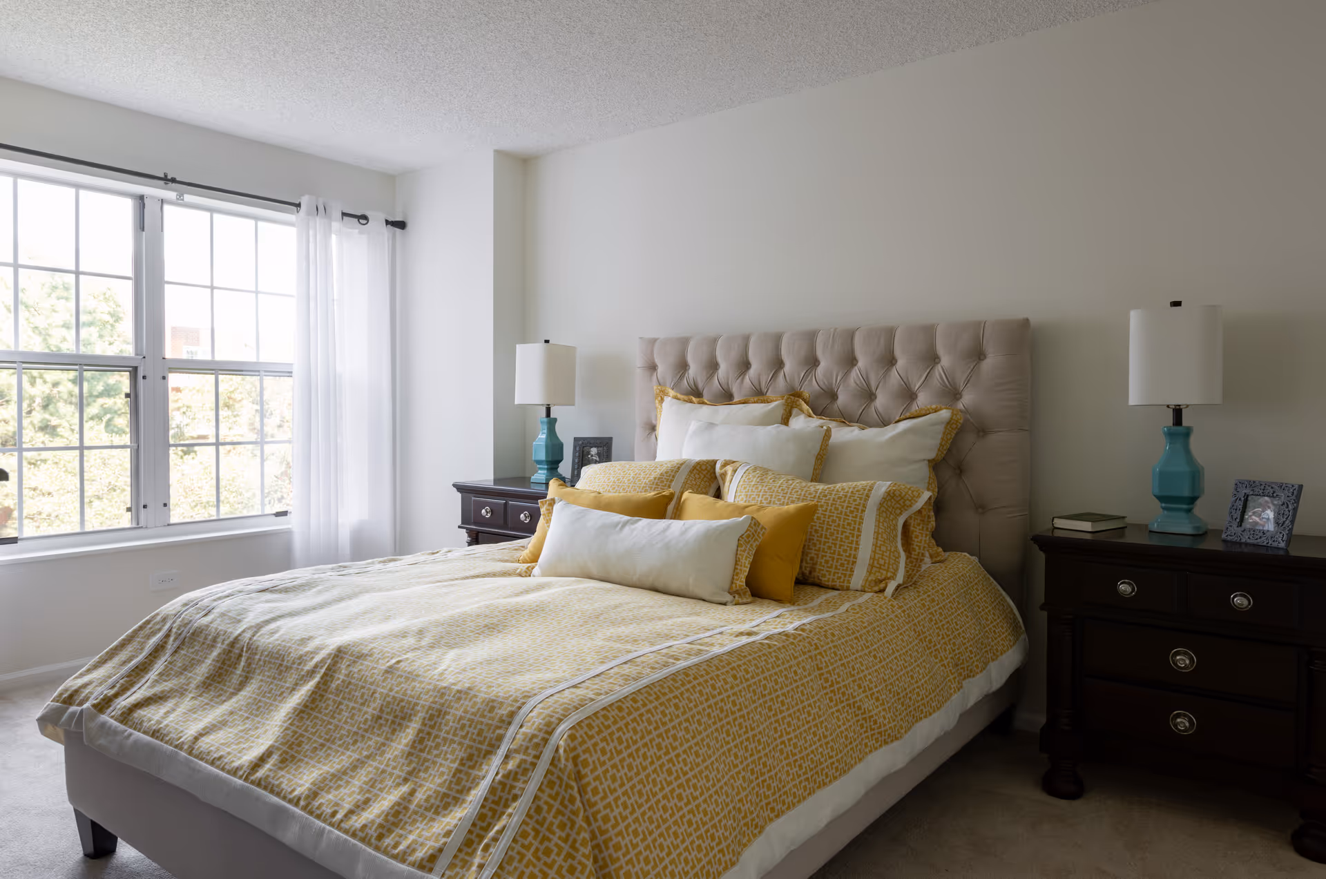A bright bedroom with a large window covered by sheer white curtains. The bed has a tufted beige headboard and is made with yellow and white patterned bedding and multiple pillows. There are two dark wooden nightstands on either side of the bed, each with a turquoise lamp and some decorative items.