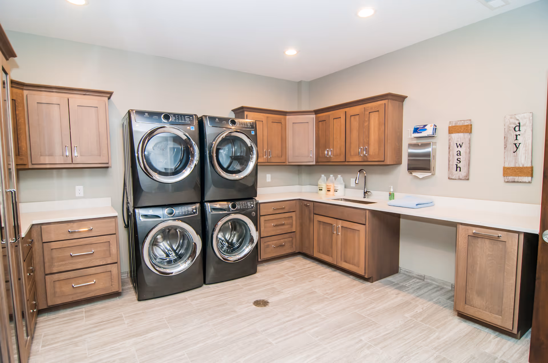 A bright laundry room with four stacked front-loading washers and dryers, wooden cabinets above and below white countertops, a sink with a faucet, and wall decorations labeled 'wash' and 'dry'.