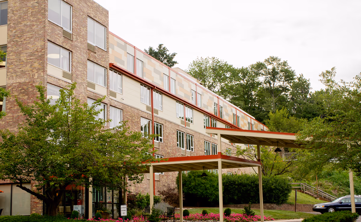 Exterior view of a multi-story brick and panel building with large windows, surrounded by trees and landscaping. There are covered walkways with red trim and a parked car visible on the right side. The sky is overcast.