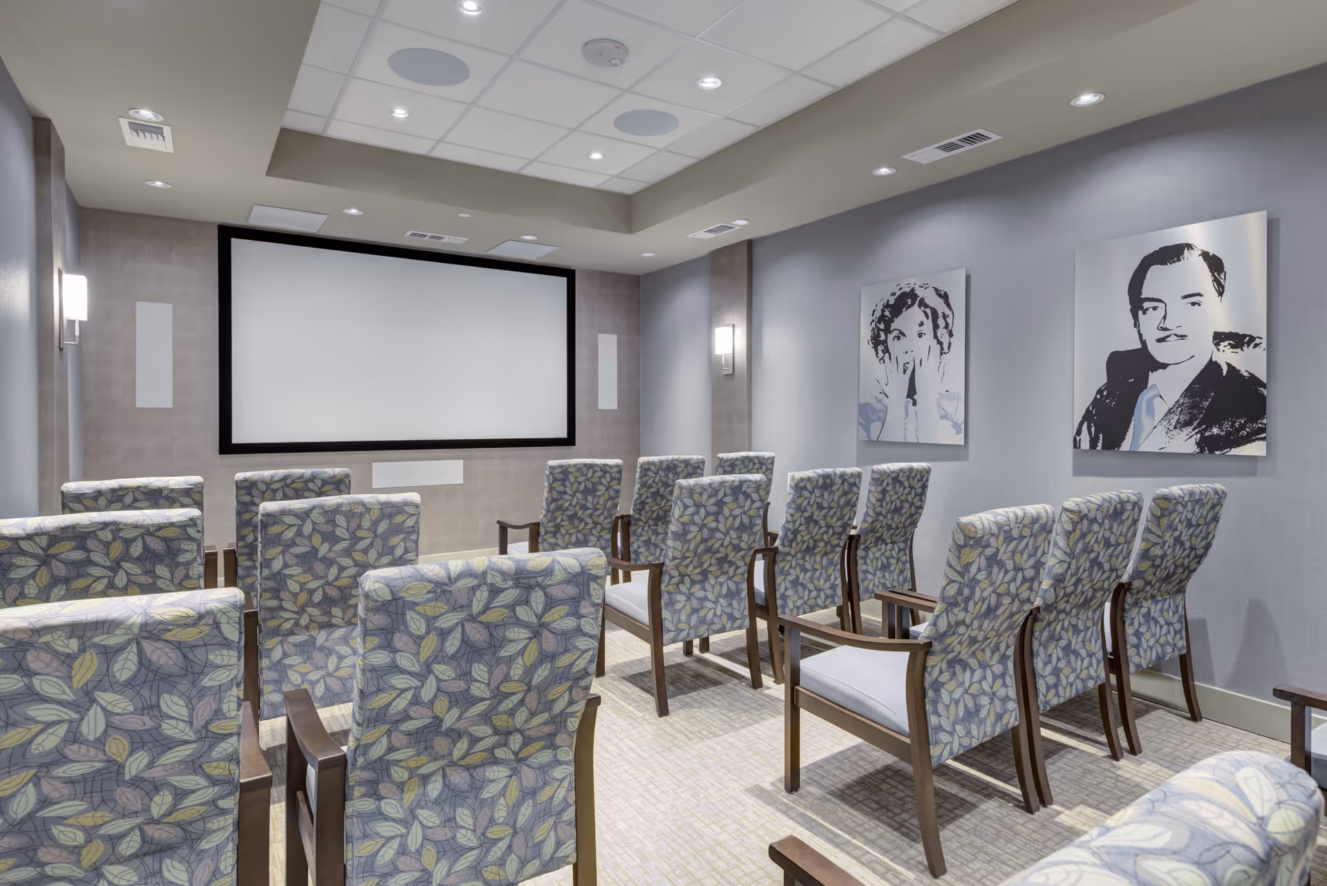 A small theater room with multiple rows of cushioned chairs featuring a leaf pattern, facing a large blank projection screen. The walls are gray with two black and white portraits hanging on the right side. The ceiling has recessed lighting and built-in speakers.