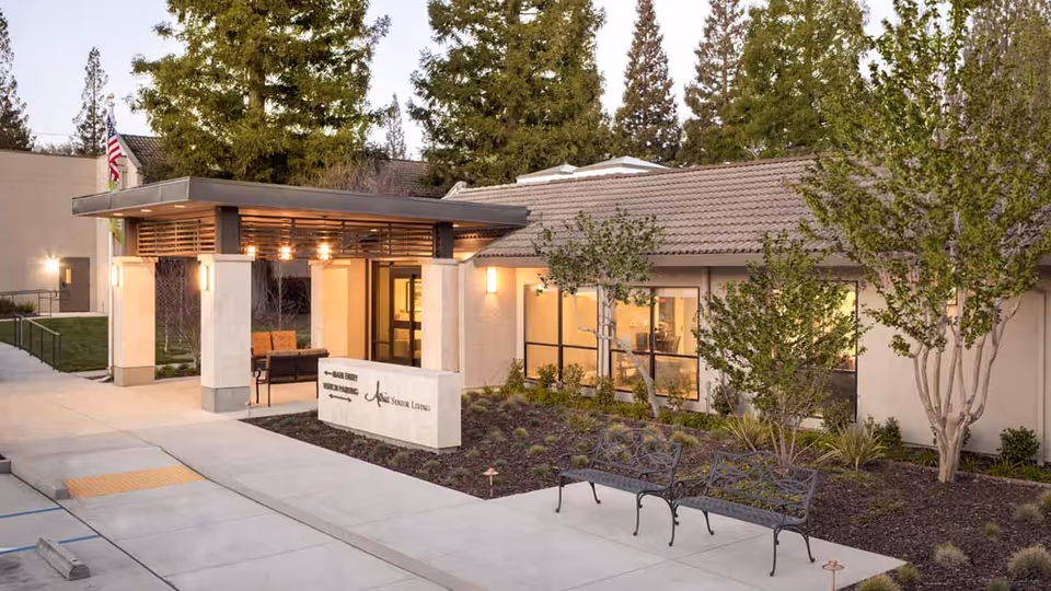 Exterior view of Atria El Camino Gardens senior living facility entrance with a covered walkway, benches, landscaped garden, and trees. The building has large windows and a tiled roof, with warm lighting inside.
