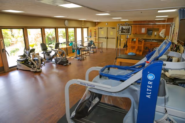 A spacious exercise room with various fitness equipment including recumbent bikes, a gravity treadmill, and other rehabilitation machines. The room has large windows letting in natural light and wooden flooring.