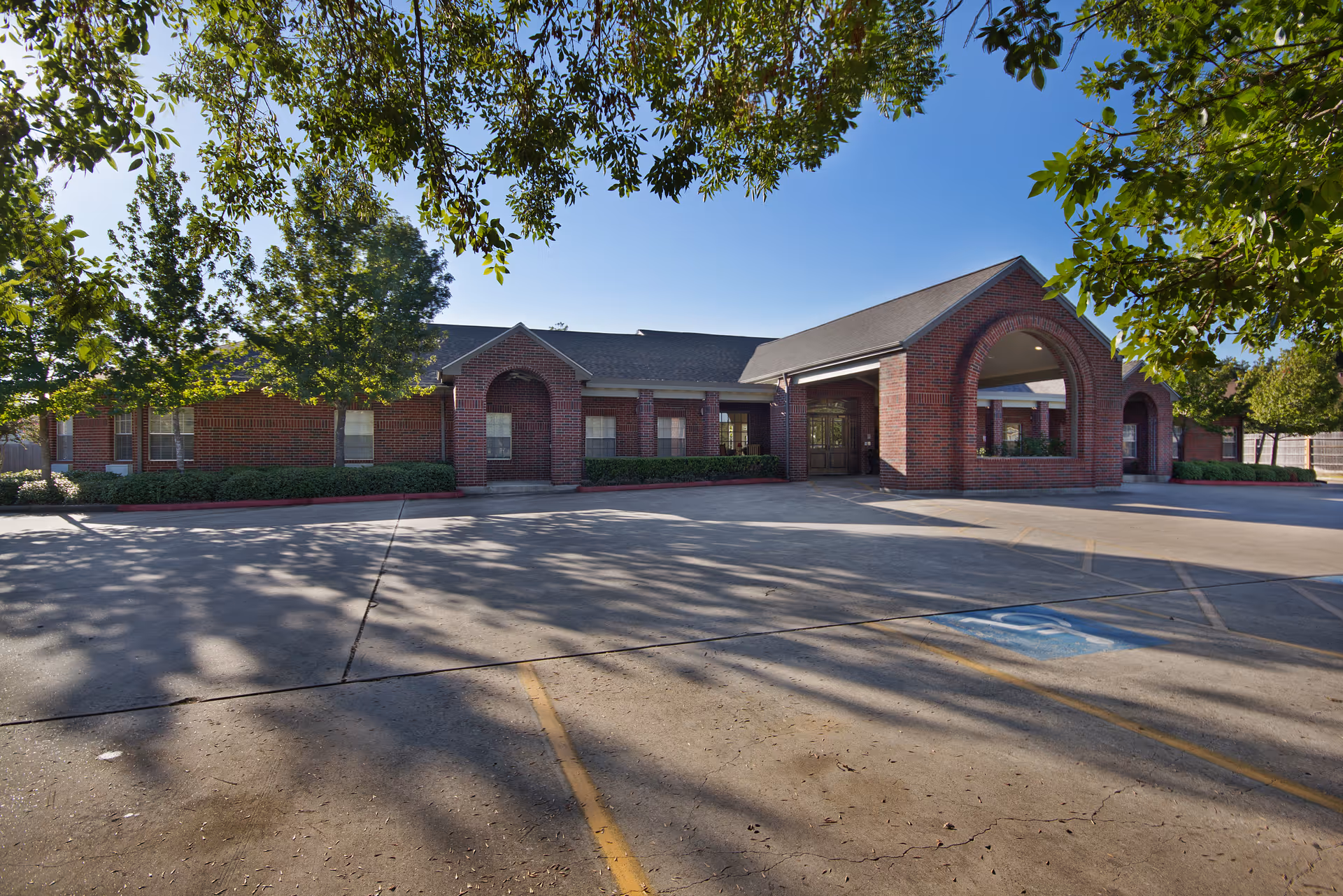 Exterior view of Braeswood Estates Senior Living facility showing a single-story brick building with an arched entrance and a covered drop-off area. The foreground features a parking lot with a designated handicapped parking space, and leafy tree branches frame the top of the image.