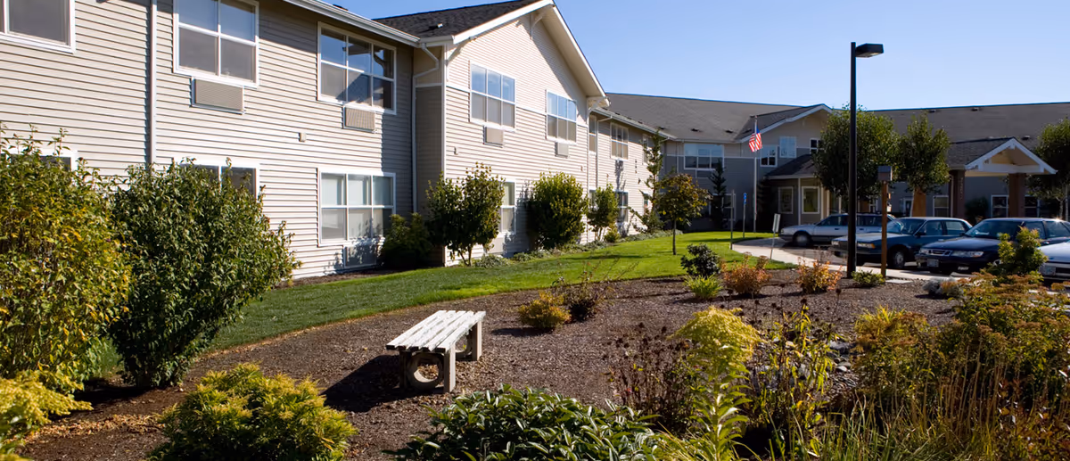 Exterior view of Avamere at Newberg senior living facility showing a two-story beige building with multiple windows, a landscaped garden with bushes and plants, a white bench, a parking area with cars, and a clear blue sky.