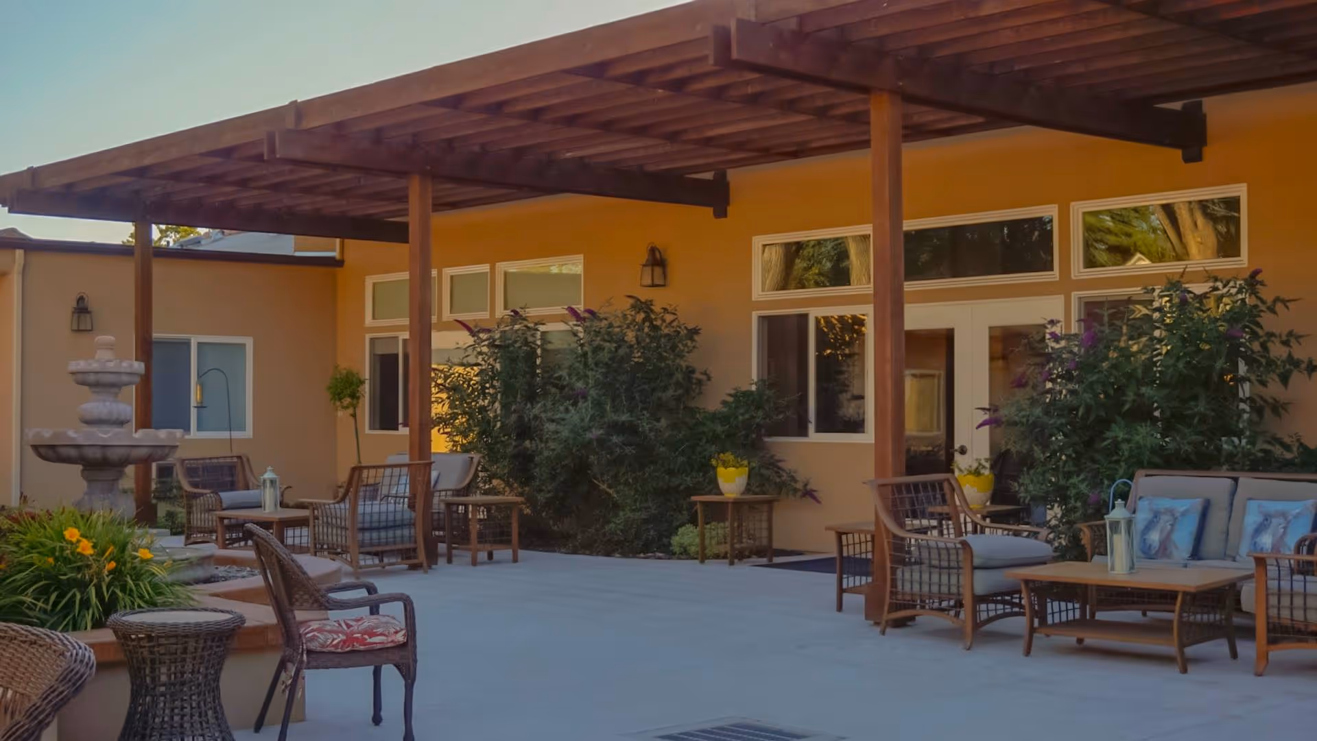 Outdoor patio area at Annette Lodge with wooden pergola, cushioned wicker chairs, tables, potted plants, and a multi-tiered stone fountain surrounded by greenery.
