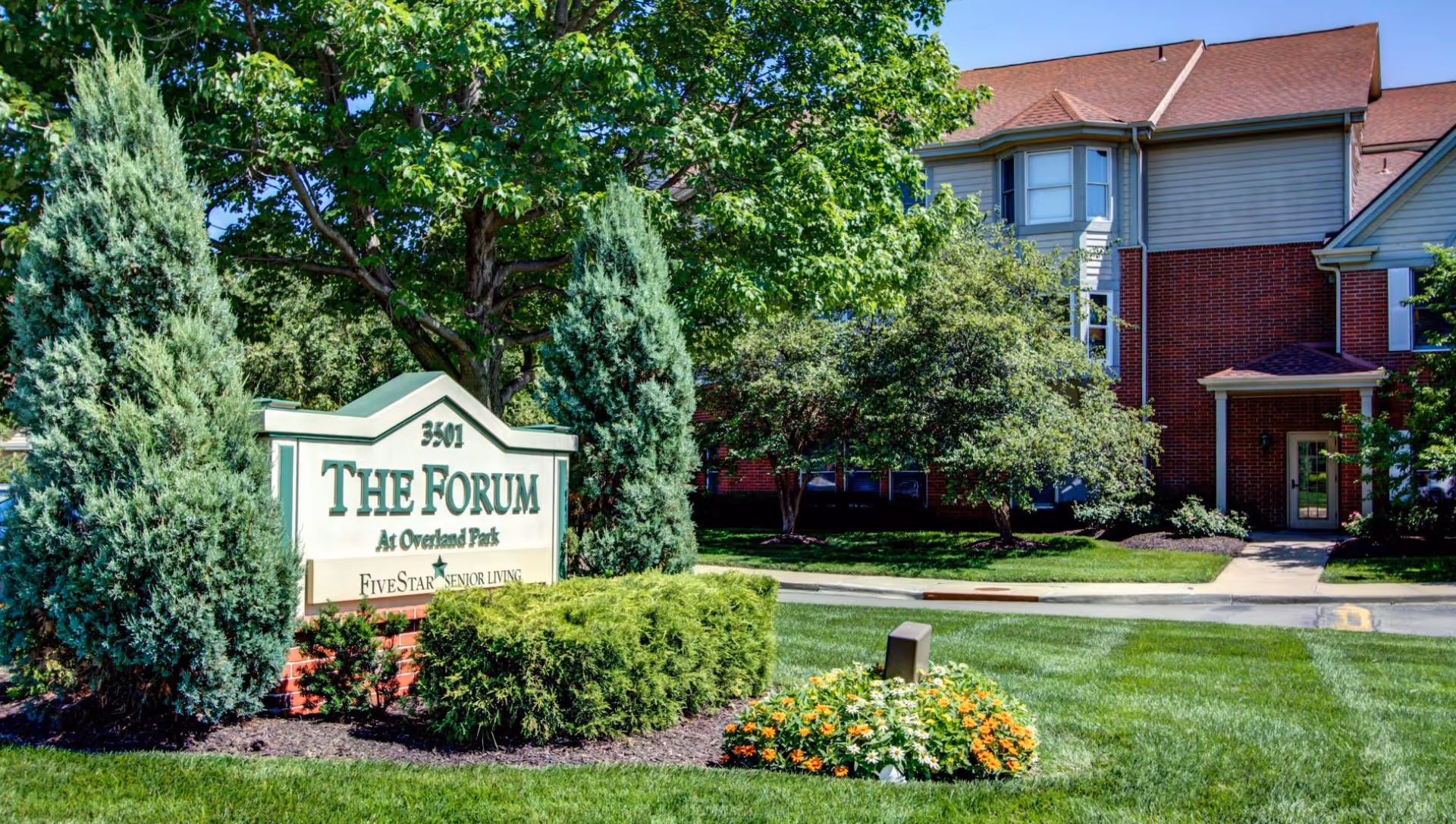 Exterior view of The Forum at Overland Park senior living facility showing a large sign with the facility name surrounded by green bushes and trees, with part of the building visible in the background under a clear blue sky.