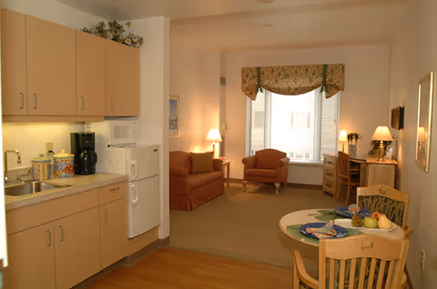 Interior view of a senior living facility apartment showing a small kitchen area with light wood cabinets, a white refrigerator, microwave, and coffee maker. Adjacent to the kitchen is a dining area with a round table set with plates and fruit. Beyond the dining area is a living room with two upholstered chairs, a small sofa, a desk with a lamp, and a window with a floral valance.