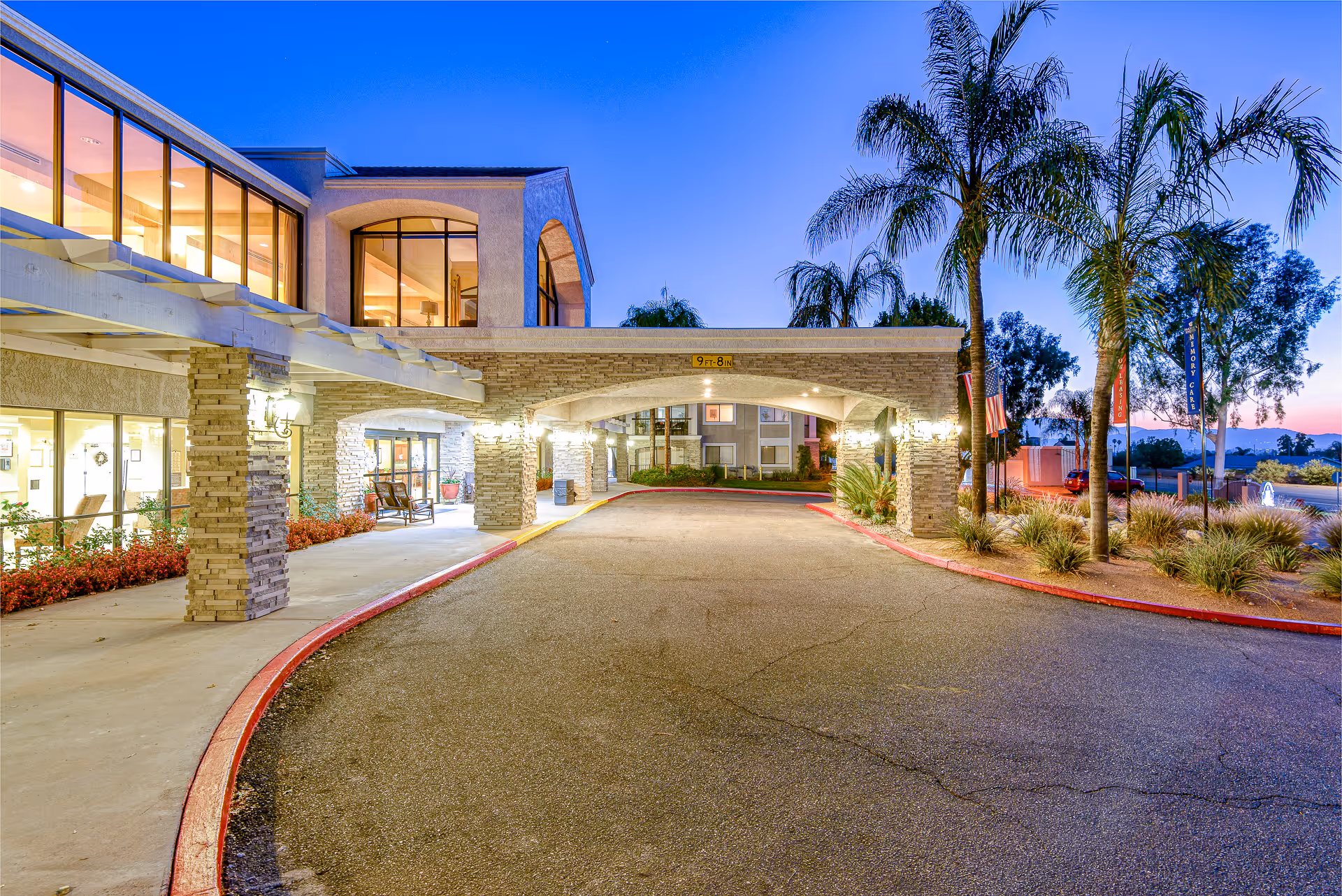 Exterior view of Menifee Senior Living facility at dusk showing the entrance driveway with stone pillars and archway, large windows with interior lights on, palm trees, and landscaping along the driveway.