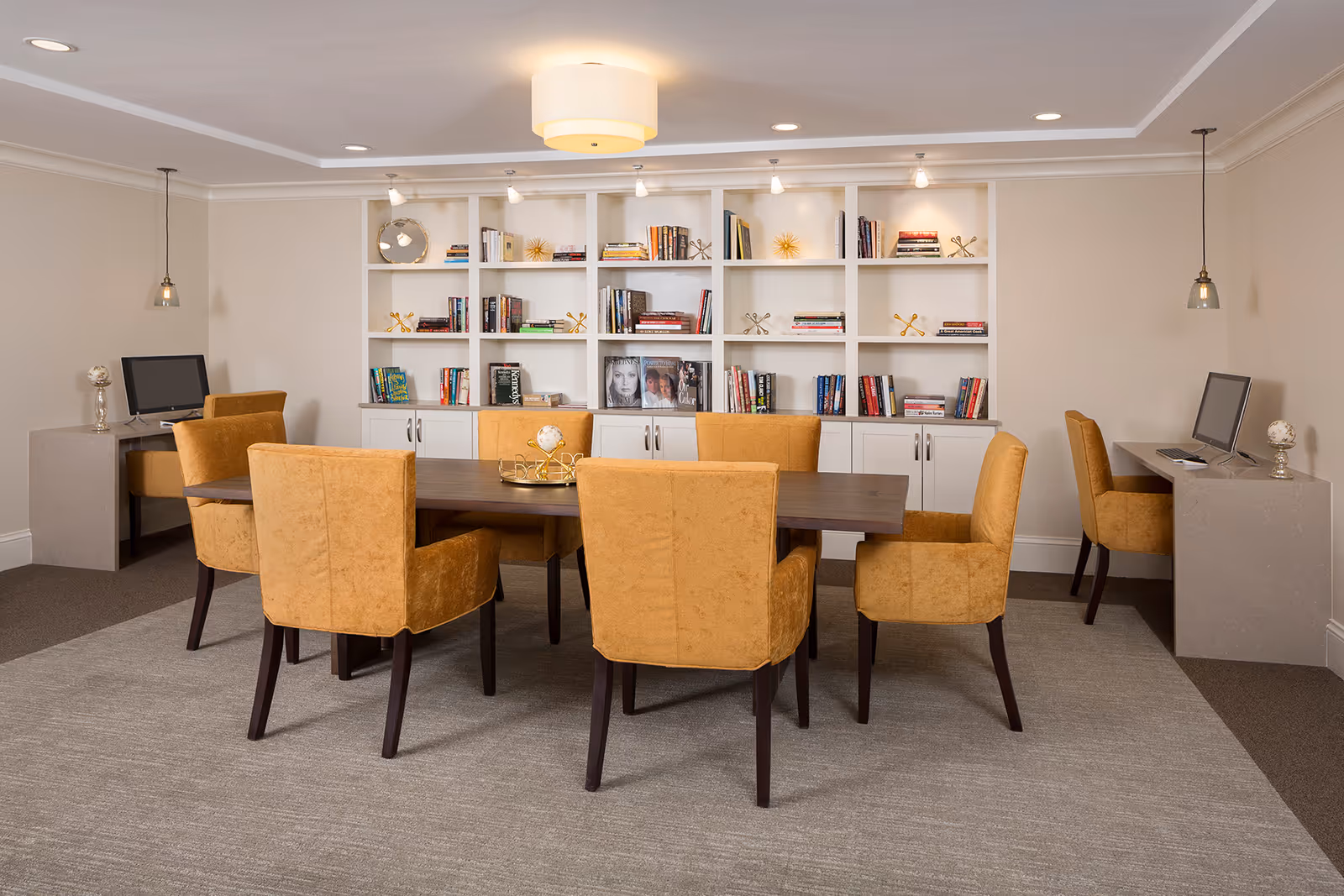 A well-lit room with a large wooden table surrounded by eight mustard-yellow upholstered chairs. Behind the table is a white built-in bookshelf filled with books and decorative items. On each side of the room, there are desks with computers and matching chairs. The room has a beige carpet and neutral-colored walls with modern ceiling lights.