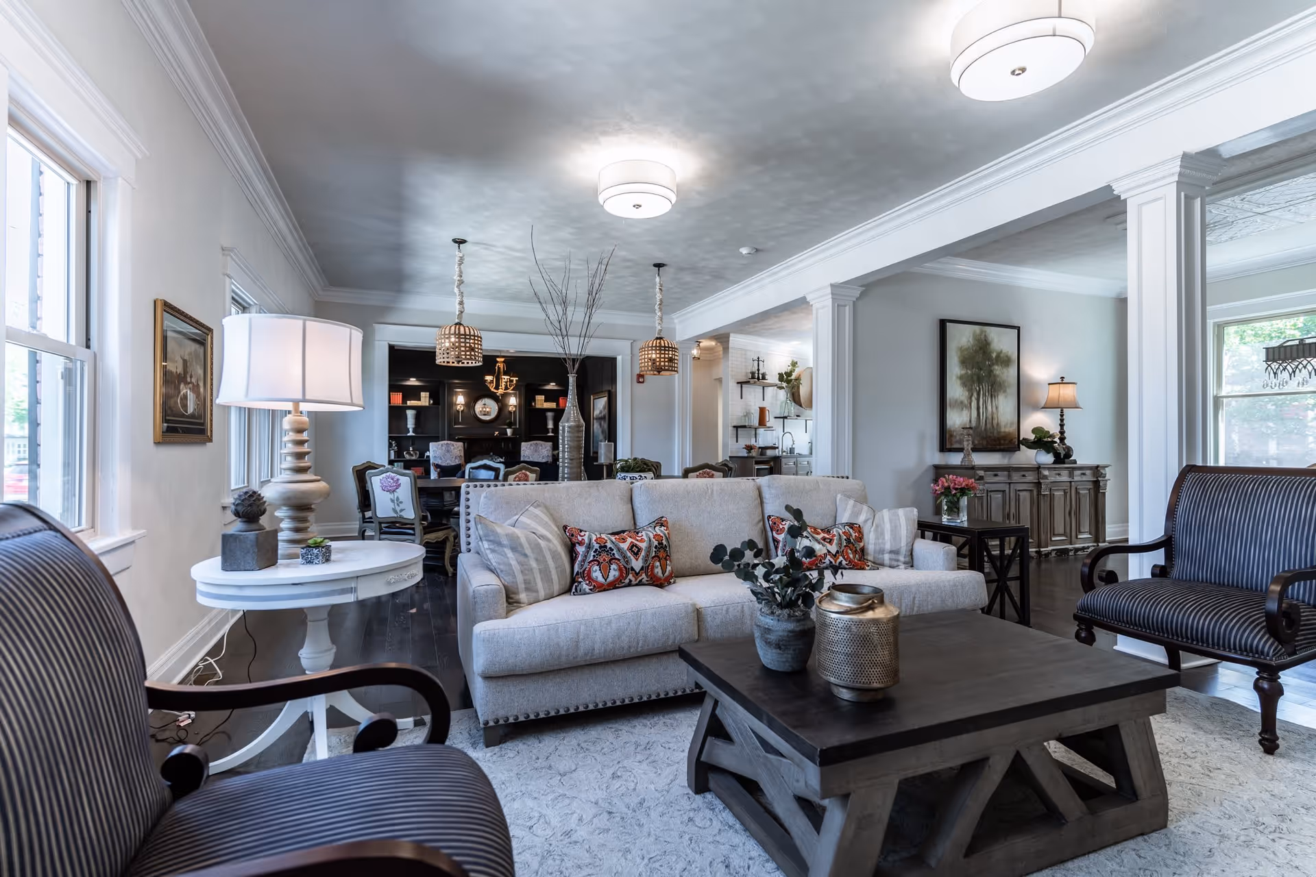 A bright and spacious living room in Elm Haven Place featuring a beige sofa with patterned pillows, two striped armchairs, a dark wooden coffee table with decorative items, a white side table with a lamp, and large windows letting in natural light. The room has white walls, crown molding, and dark hardwood floors. In the background, there is a dining area with a table and chairs, pendant lights, and built-in shelving.