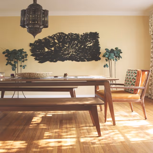 A dining room with a wooden table and bench, a wooden chair with a patterned cushion, two potted plants, a decorative black wall art piece, and a hanging lantern-style light fixture. Sunlight casts shadows on the wooden floor.