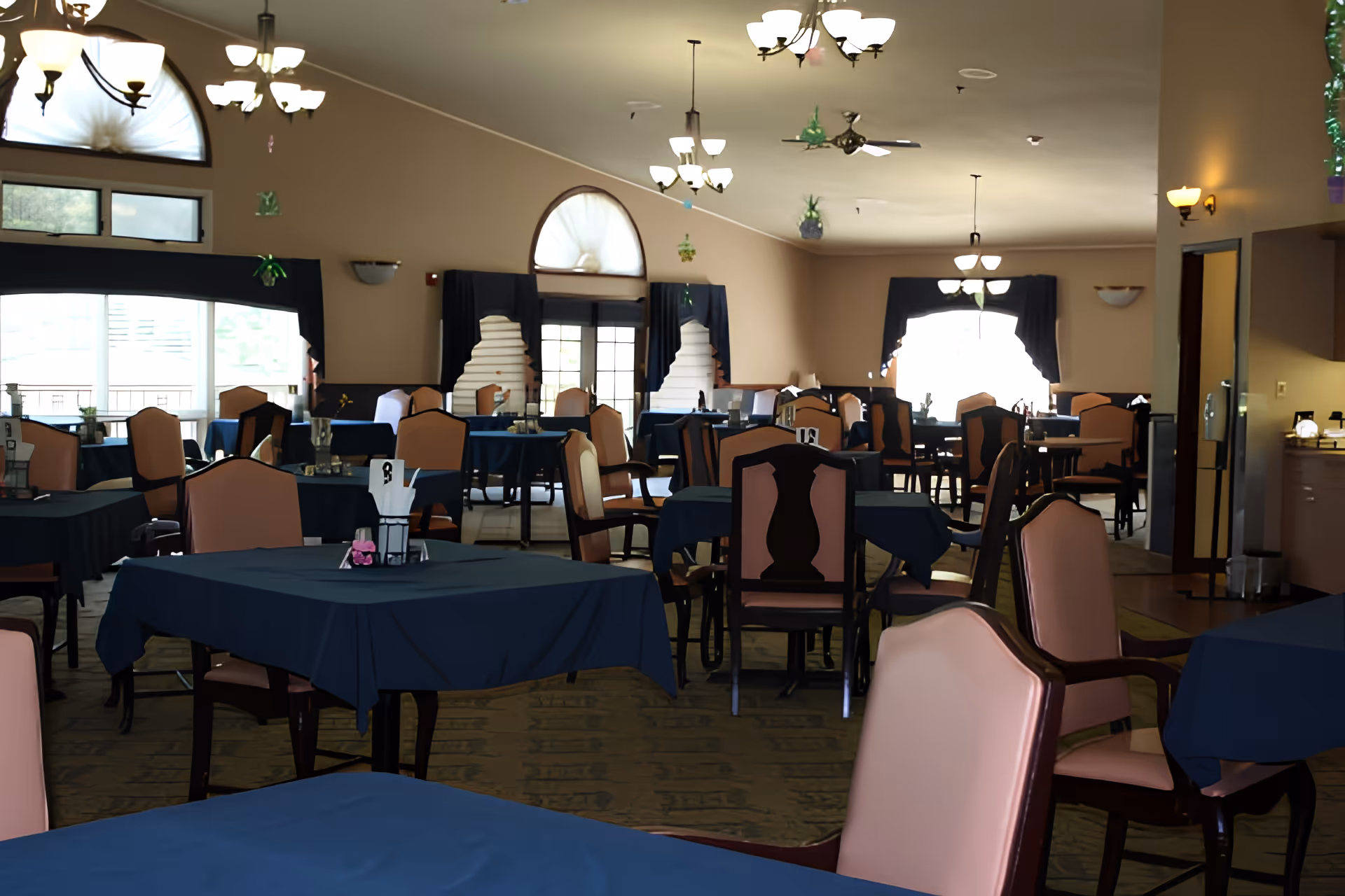Interior view of a senior living center dining room with multiple tables covered in dark blue tablecloths and surrounded by cushioned chairs with wooden frames. The room has large windows with arched tops and dark curtains, and several ceiling light fixtures providing warm lighting. Some tables have small decorative items and napkin holders.