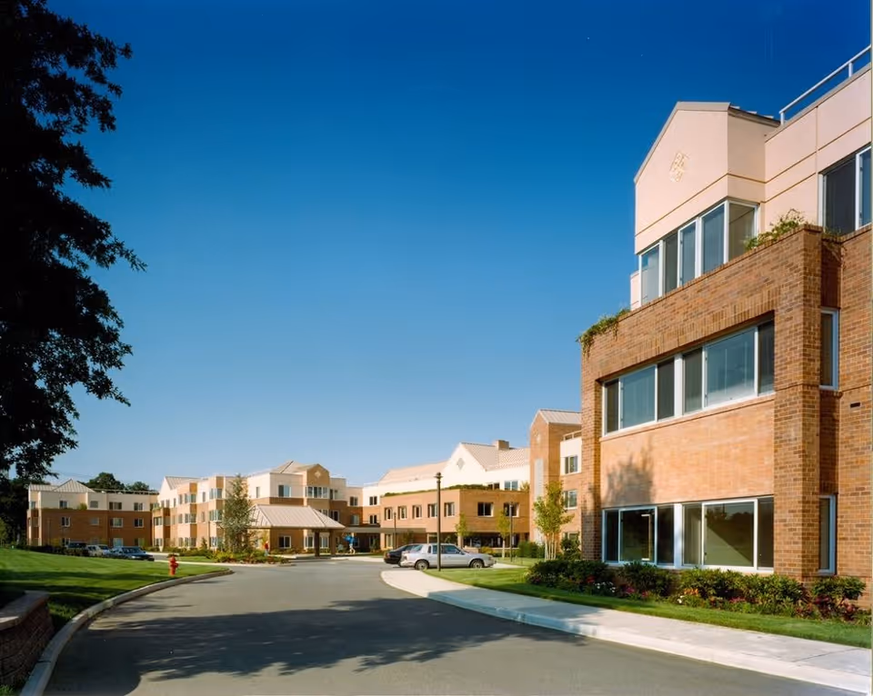 Exterior view of Gurwin Jewish Nursing & Rehabilitation Center showing a multi-story brick building with large windows, a driveway, parked cars, and landscaped greenery under a clear blue sky.