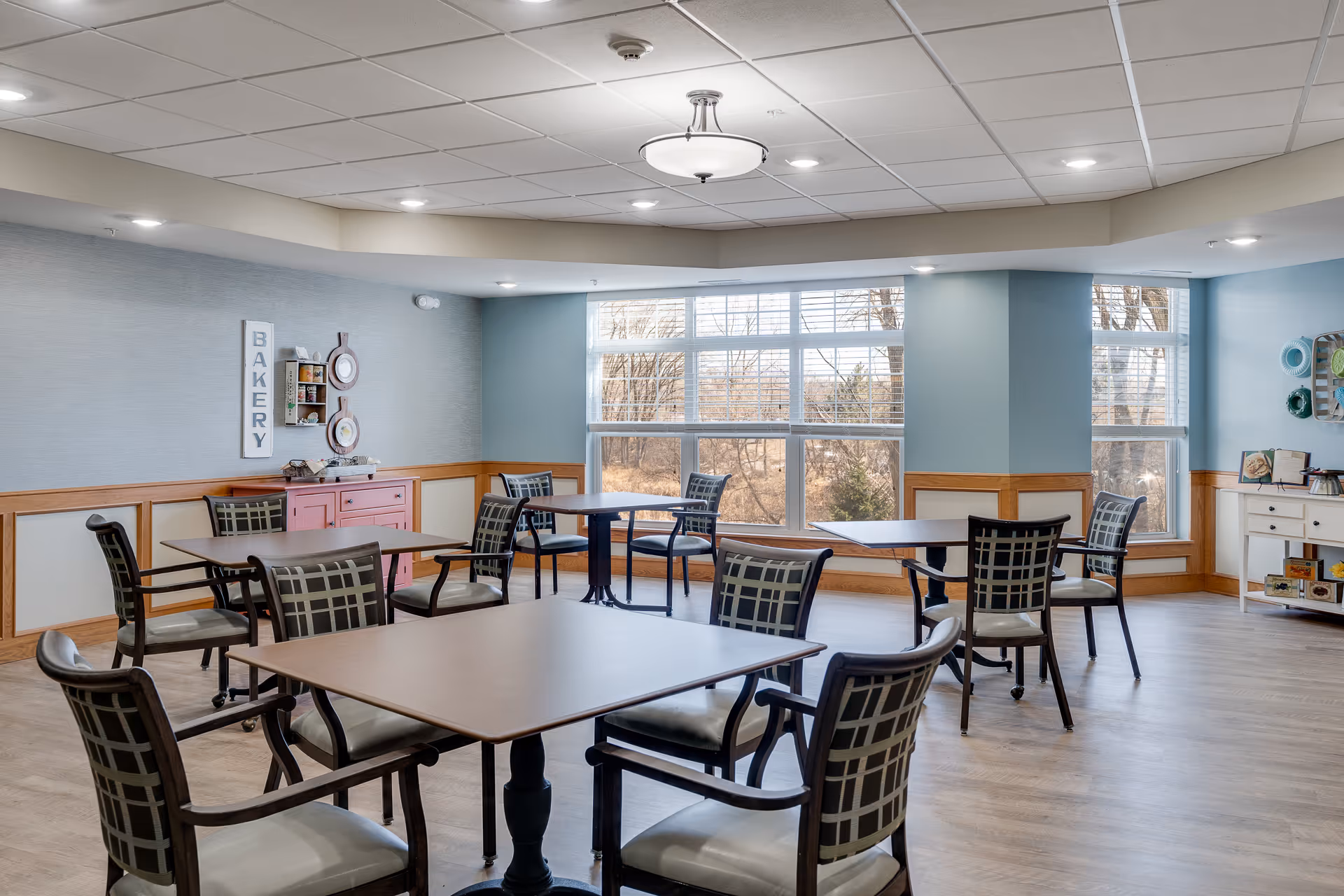 A bright dining room with multiple square tables and cushioned chairs arranged neatly. Large windows allow natural light to fill the room, showing a view of trees outside. The walls are painted in soft blue and beige tones with wooden paneling. Decorative items, including a sign that says 'BAKERY' and colorful plates, adorn the walls. The floor is light wood, and ceiling lights provide additional illumination.