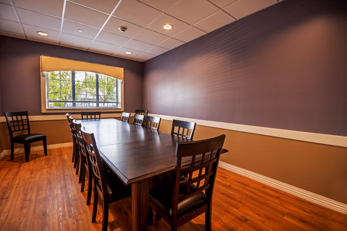 A dining room with a long dark wooden table surrounded by ten matching chairs. The room has wooden flooring, two-tone walls with a beige lower half and purple upper half, and a large window with a beige valance letting in natural light.