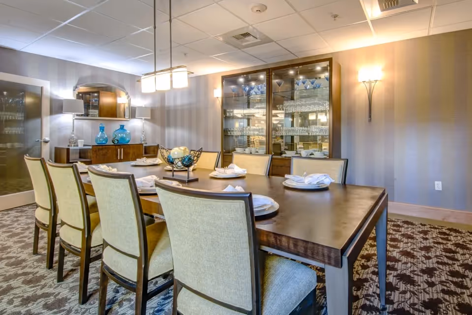 A formal dining room with a large wooden table set for six people with plates and napkins. The room features upholstered chairs, a patterned carpet, a glass-front cabinet filled with glassware, and a sideboard with decorative blue vases and lamps. The walls have a subtle striped pattern and are lit by wall sconces and a hanging light fixture above the table.