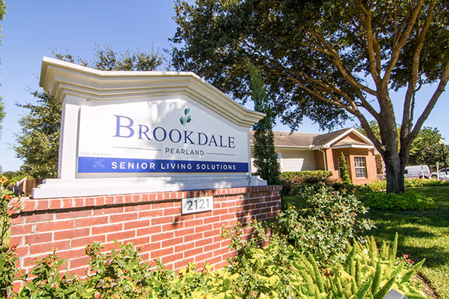 Outdoor view of the entrance sign for Brookdale Pearland Senior Living Solutions, featuring a brick base with greenery and trees around, and a building partially visible in the background under a clear blue sky.