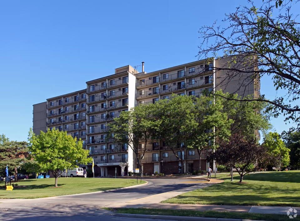 Exterior view of a multi-story residential building named Alpha Towers surrounded by green trees and a well-maintained lawn under a clear blue sky.
