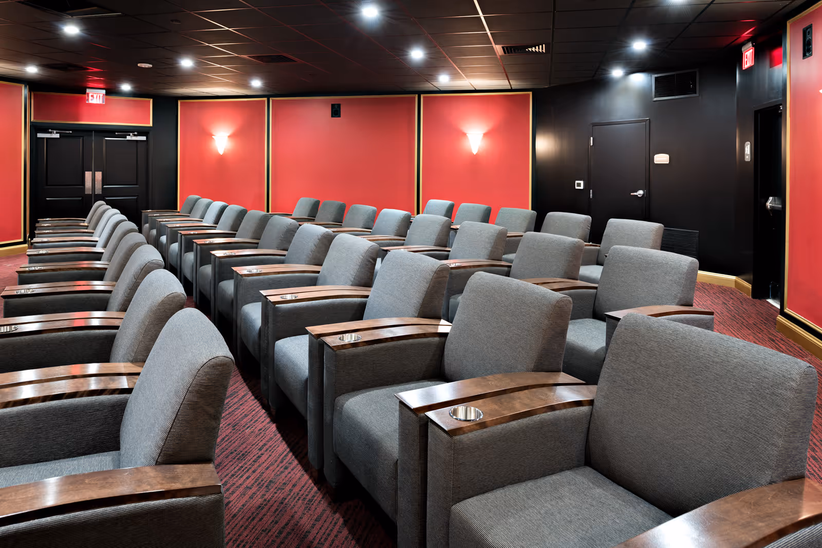Interior view of a small theater or screening room with multiple rows of gray upholstered chairs featuring wooden armrests with cup holders. The walls are painted red with black trim and have wall sconces providing soft lighting. The ceiling is black with recessed lighting, and there are double doors at the back of the room.