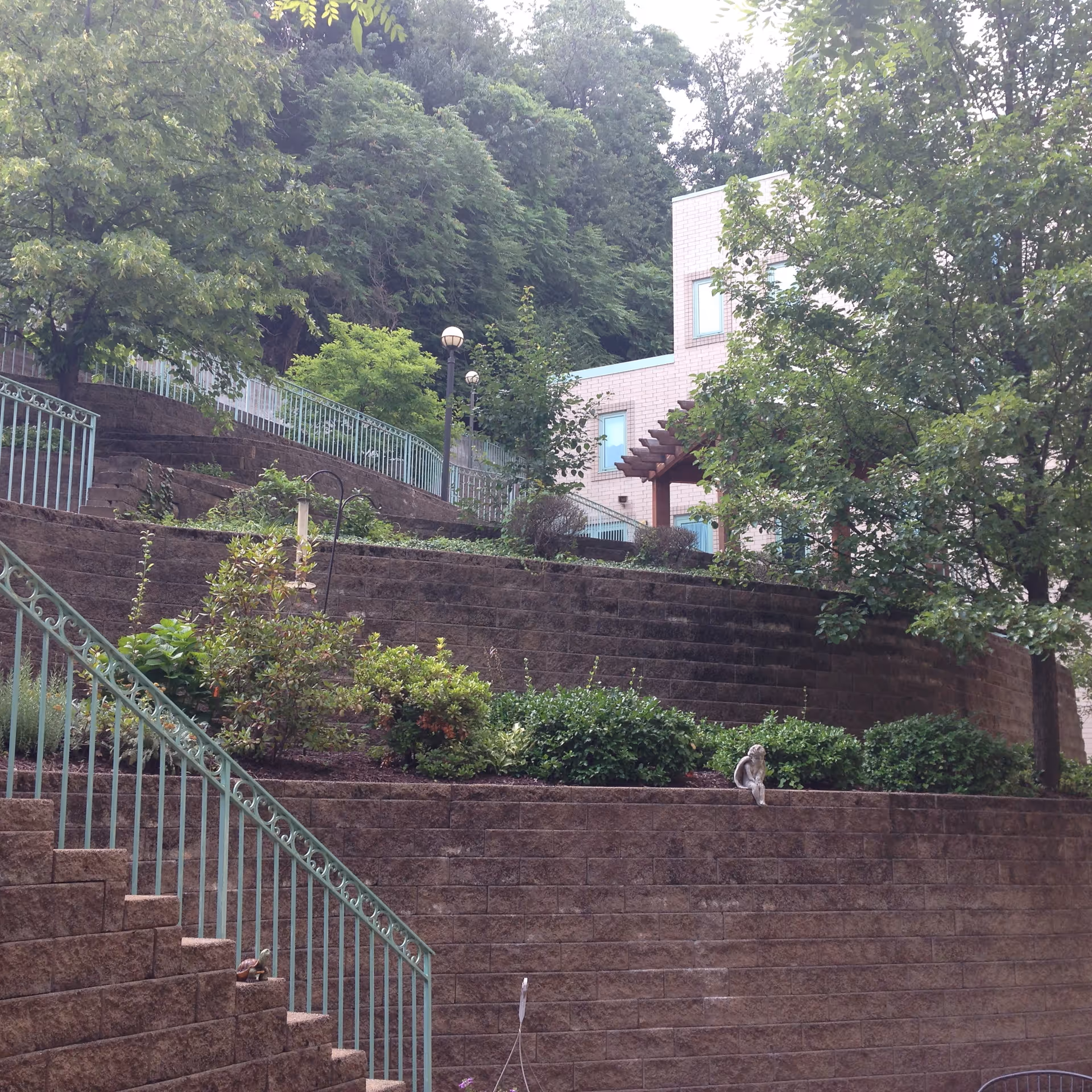 Terraced garden area with stone retaining walls and green metal railings. There are various shrubs and trees planted on the terraces, and a building with light-colored brick and windows is visible in the background. A small statue of a sitting figure is placed on one of the retaining walls.