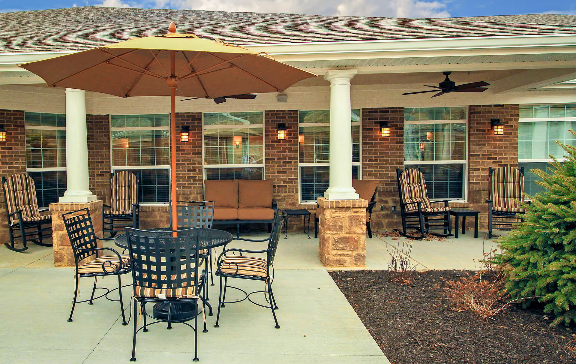 Outdoor patio area with a round metal table and four chairs with striped cushions under a large beige umbrella. Behind the table are several cushioned rocking chairs and a cushioned bench against a brick wall with windows and white columns supporting the roof. Ceiling fans are mounted under the covered patio.