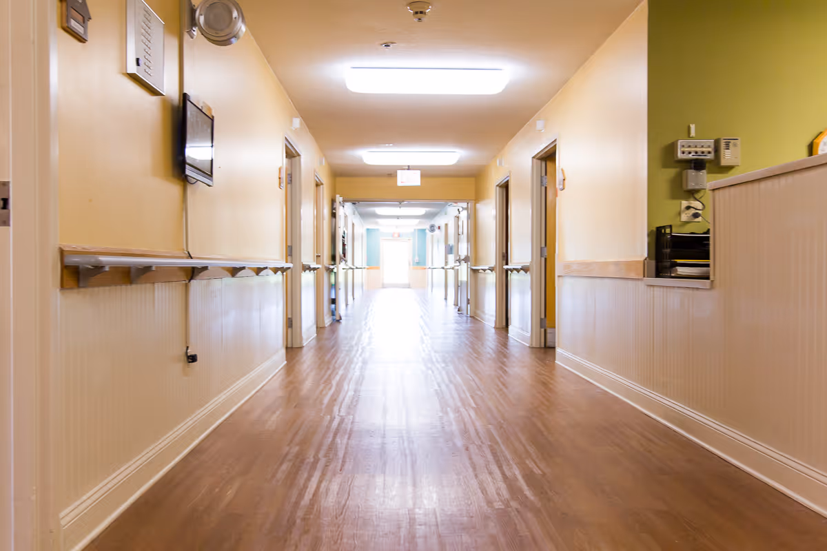 A long, well-lit hallway in a skilled nursing and rehab facility with wooden floors, beige walls, handrails along both sides, and several open doorways leading to rooms.