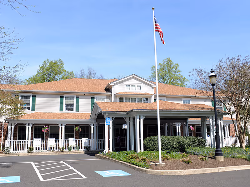 Front exterior view of a two-story senior living facility building with a covered entrance, white railings, green shutters, and a flagpole with an American flag. There are parking spaces including handicapped spots in front, and some trees and shrubs around the building.