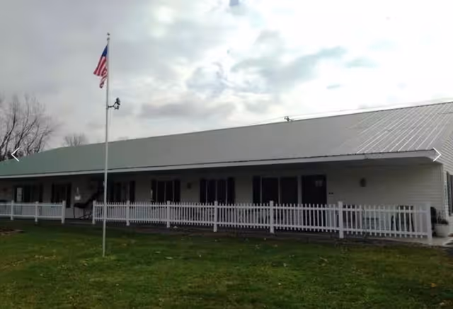 Single-story building with a white exterior and a metal roof, surrounded by a white picket fence. An American flag is flying on a flagpole in front of the building, with a grassy lawn in the foreground and a cloudy sky above.