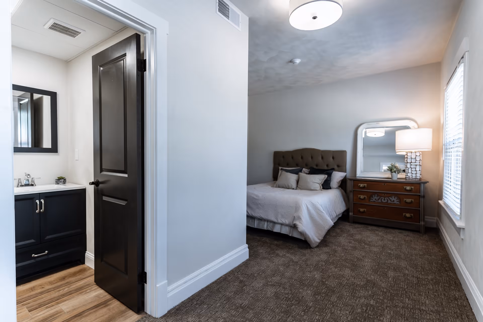 A bedroom with a bed featuring a tufted headboard and several pillows. Next to the bed is a wooden dresser with a large mirror and a table lamp. The room has carpeted flooring and a window with blinds. To the left, there is an open door leading to a bathroom with a black vanity and a mirror above the sink.