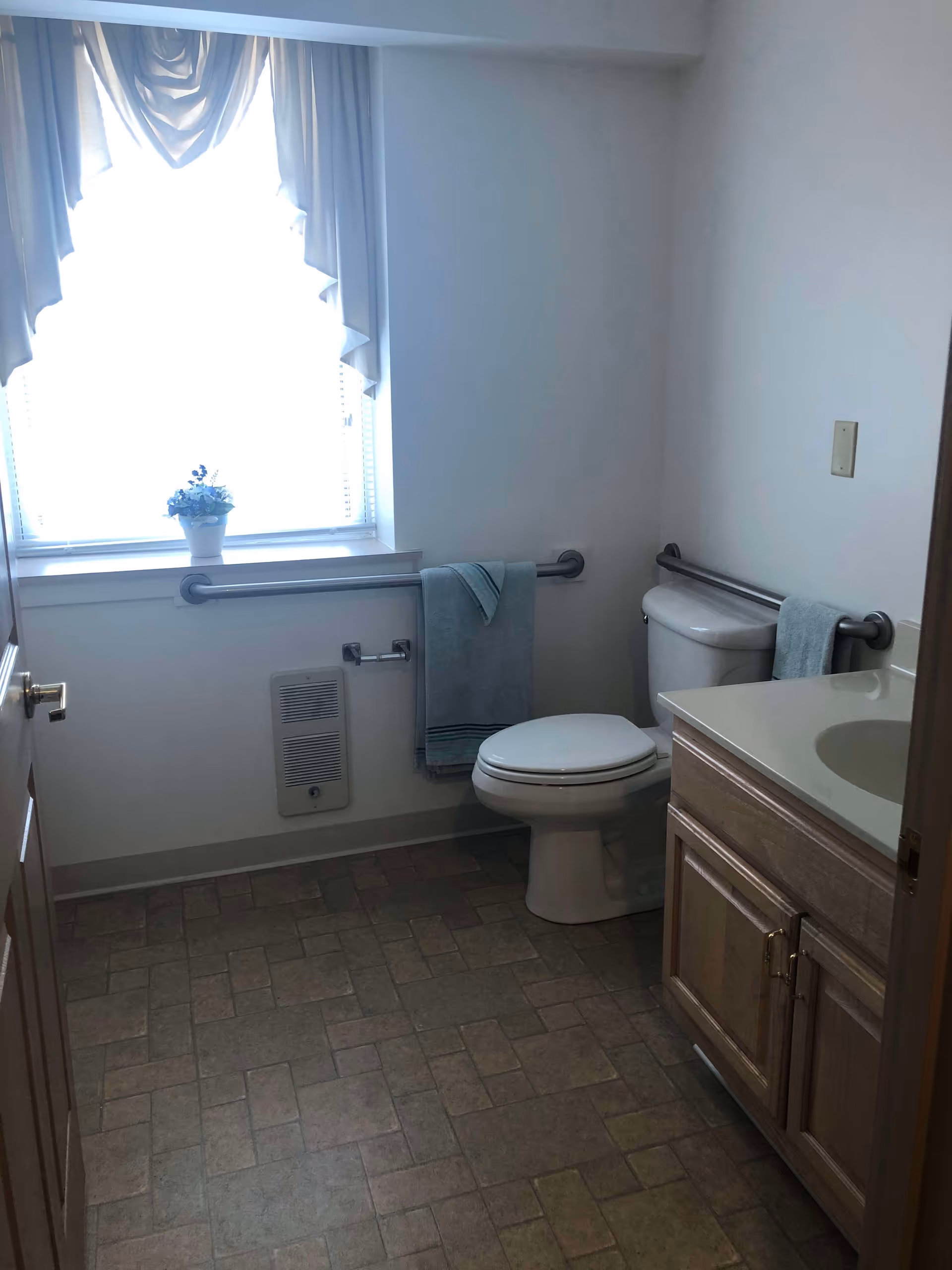 A bathroom with a window covered by white curtains, a white toilet with grab bars on both sides, a wooden vanity with a sink, and a towel hanging on a grab bar. The floor has a patterned tile design.