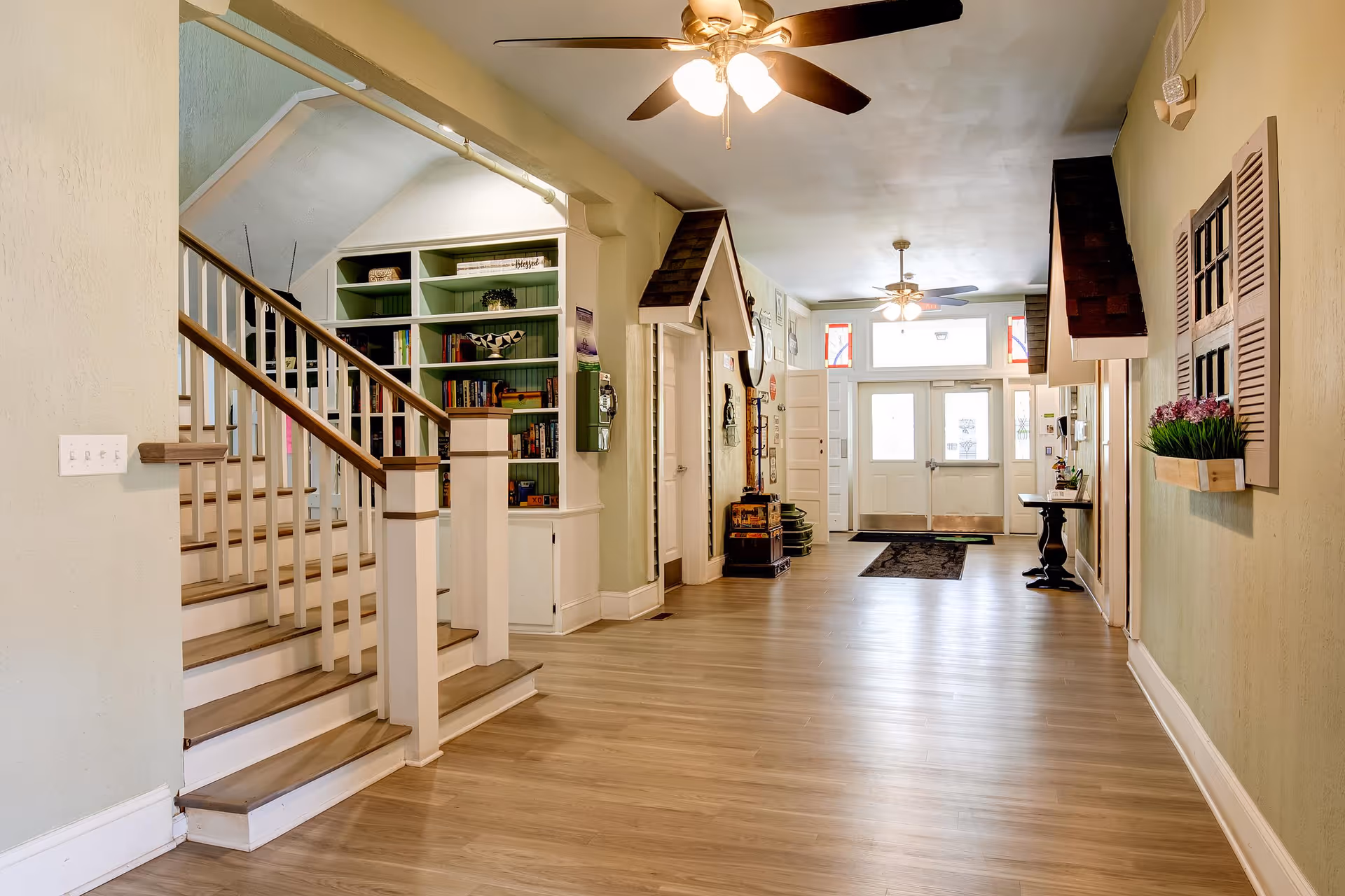Interior hallway of Shepherd Premier Senior Living featuring wooden flooring, a staircase with white railings and wooden handrails on the left, built-in green bookshelves, ceiling fans with lights, decorative wall elements including a window frame with flower box, and a double door entrance with sidelights and transom windows.