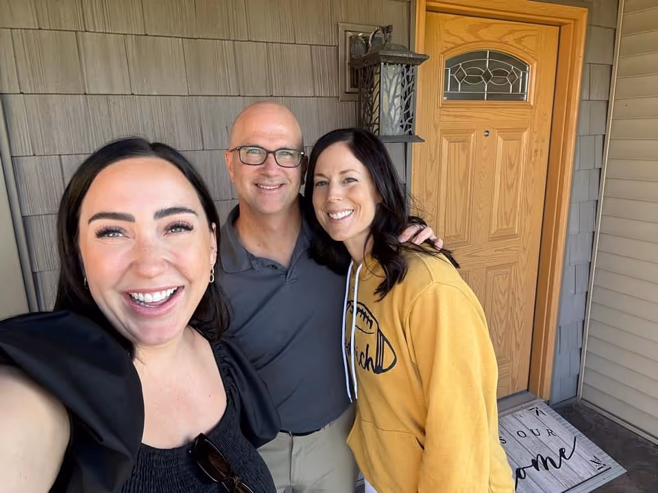 Three people smiling and posing for a selfie in front of a wooden door with a decorative glass window. The person on the left is a woman with dark hair wearing a black top, the person in the middle is a bald man wearing glasses and a gray polo shirt, and the person on the right is a woman with dark hair wearing a yellow hoodie. The setting appears to be the entrance of a house or facility with gray siding and a welcome mat on the floor.