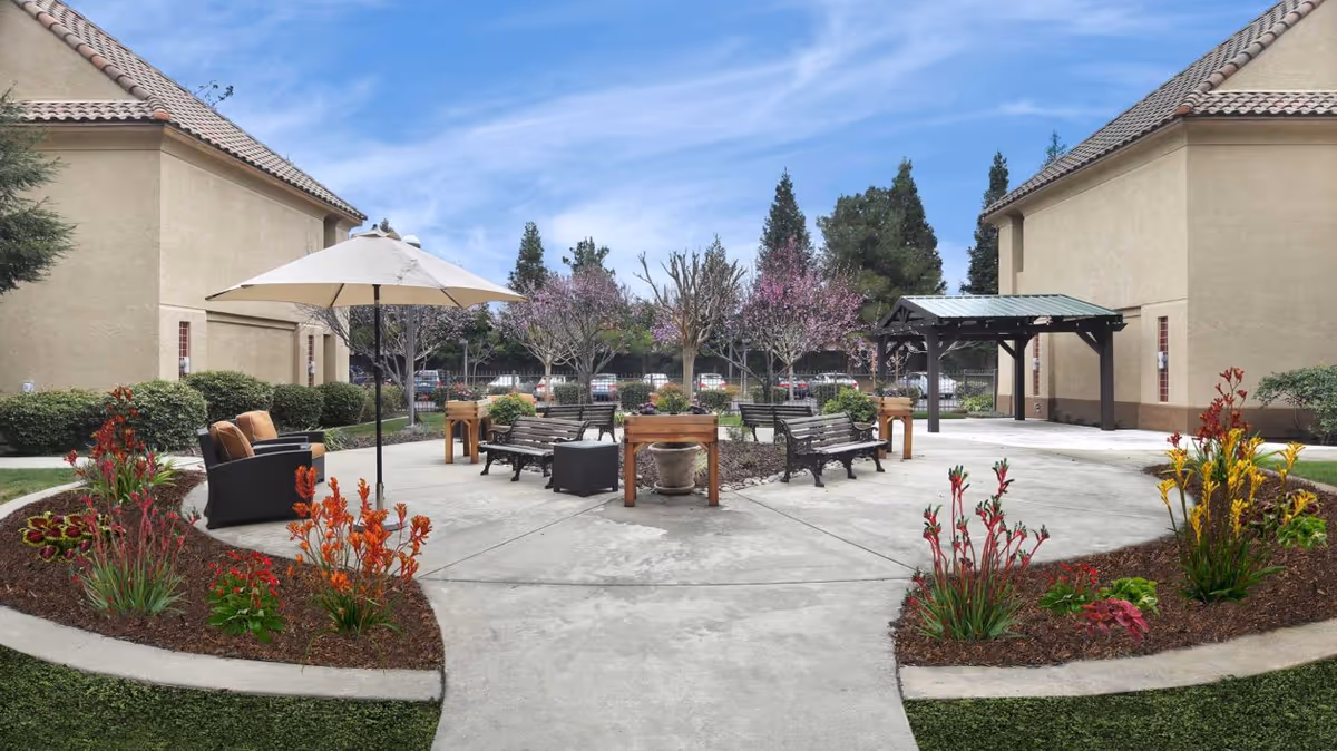 Outdoor courtyard area at Ivy Park at Roseville featuring benches, a patio umbrella with seating, flower beds with colorful plants, and trees with pink blossoms, surrounded by two beige buildings with tiled roofs under a blue sky.