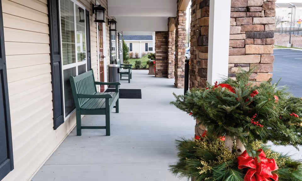 Covered outdoor walkway at a senior living facility with green benches along the wall, stone pillars, and festive holiday greenery decorations with red ribbons. A parking lot and other buildings are visible in the background.