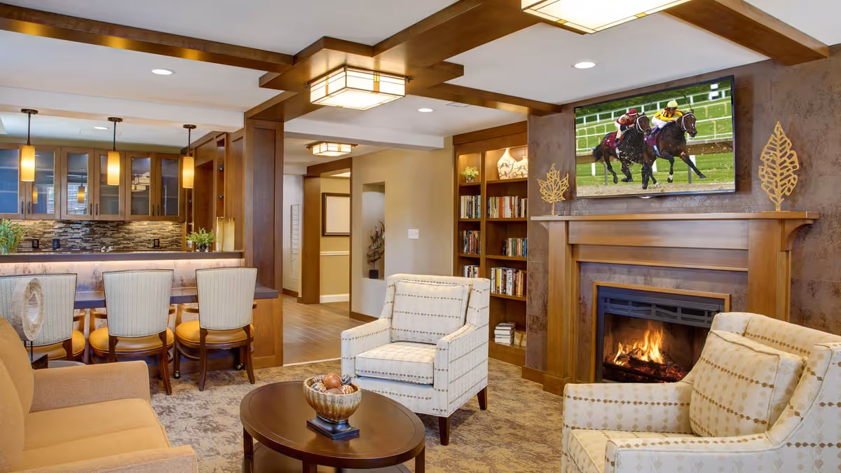 A cozy living room area in a senior living facility featuring a fireplace with a mounted TV showing a horse race. The room has two patterned armchairs, a beige sofa, a round wooden coffee table with a decorative bowl, and a built-in bookshelf. In the background, there is a kitchen area with a bar counter and four high chairs, pendant lighting, and wooden cabinetry.