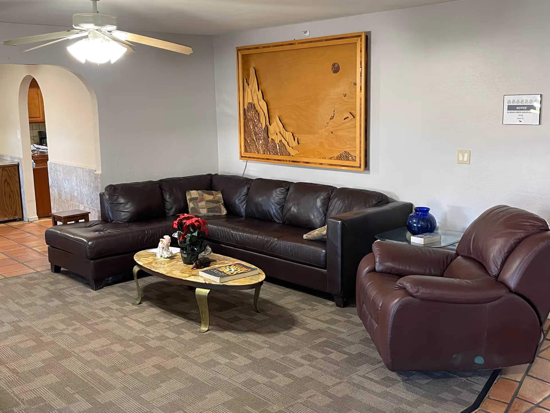 A cozy living room area with a dark brown leather sectional sofa and a matching leather armchair. A small oval coffee table with a marble top sits in front of the sofa, decorated with a red flower arrangement and a few small items. On the wall above the sofa is a large wooden artwork. The floor is carpeted with a patterned design, and there is a tiled floor area visible leading to an arched doorway into another room. A small side table with a blue vase and a book is next to the armchair.