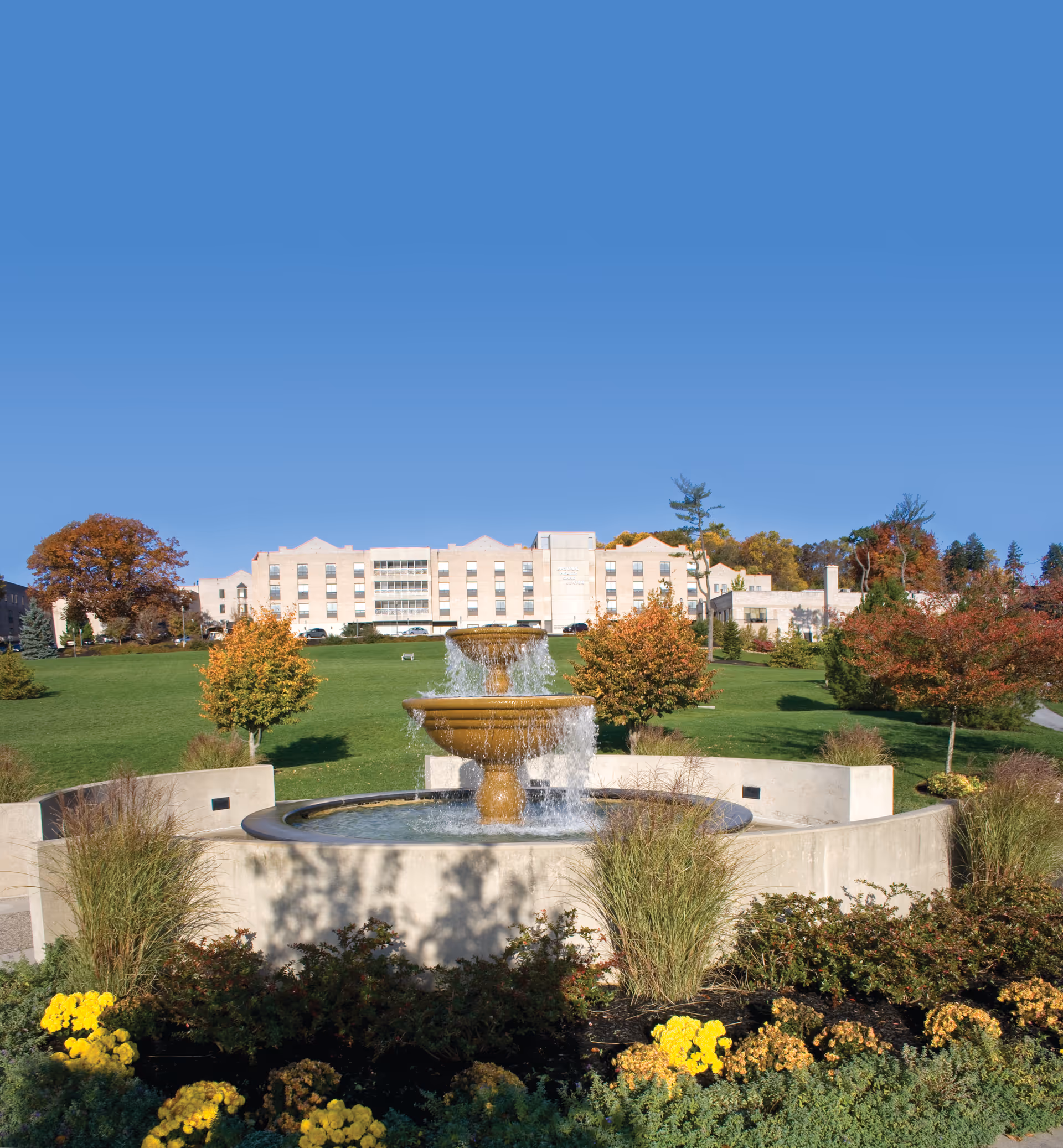 A large, multi-story beige building in the background with a green lawn and colorful autumn trees in front. In the foreground, there is a tiered water fountain surrounded by a circular concrete structure and landscaped with bushes and yellow flowers under a clear blue sky.