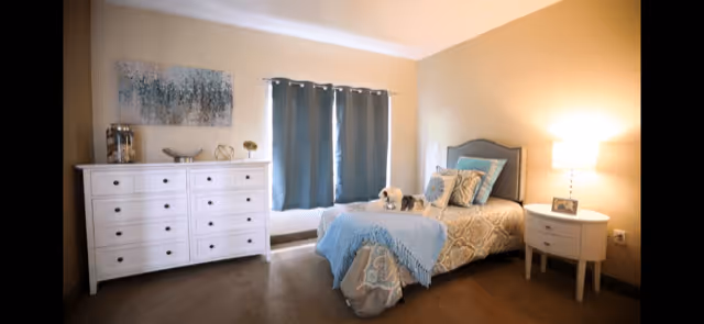 A cozy bedroom with a single bed featuring patterned bedding and multiple pillows. Next to the bed is a round white nightstand with a lamp and a small framed photo. Across from the bed is a white dresser with decorative items on top, and a window with blue curtains in the center of the wall. The walls are painted a warm beige color.