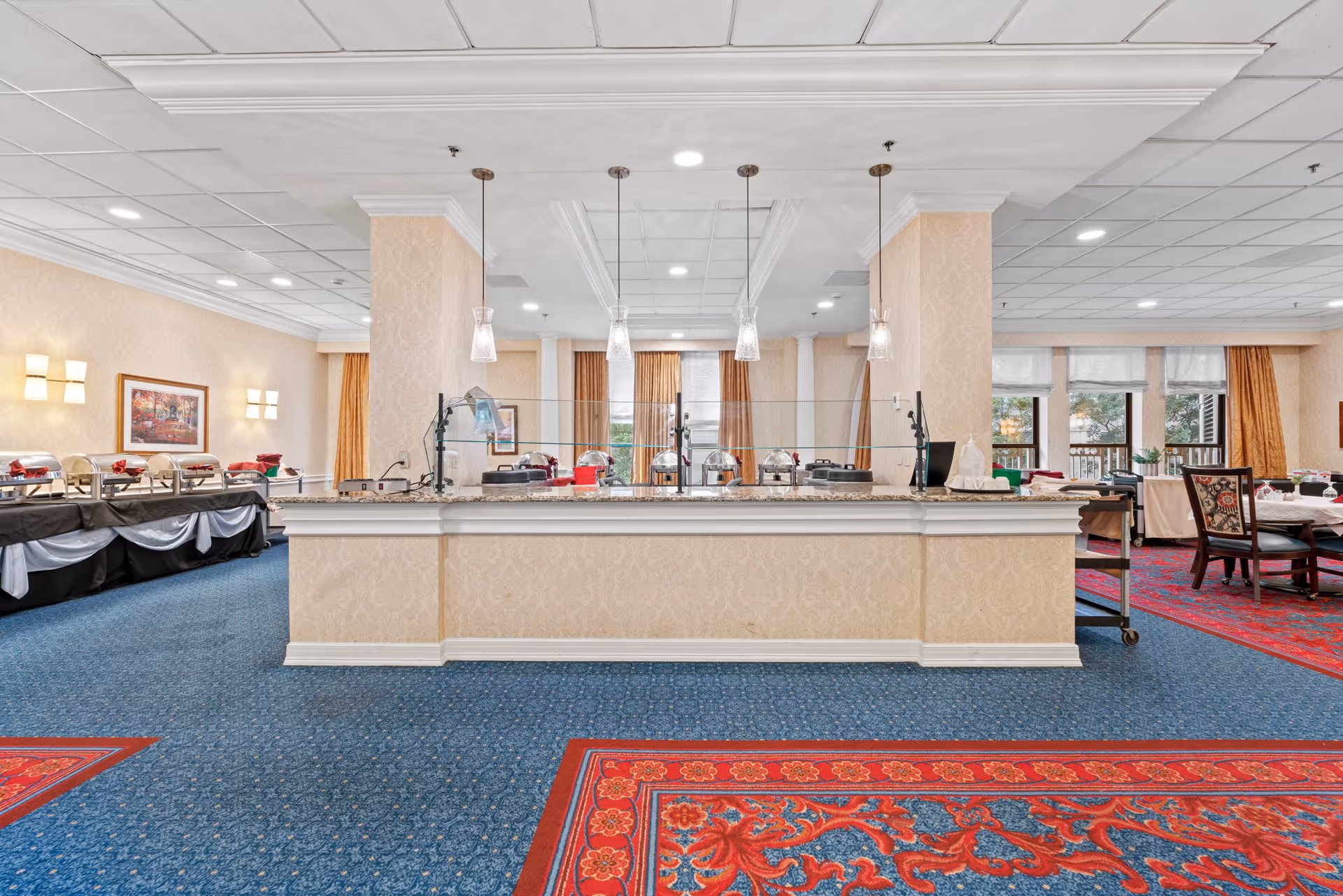 Interior view of a dining area in a senior living facility with a buffet counter in the center, covered food trays on the left side, and dining tables with chairs on the right. The room has blue carpet with red patterned rugs, beige walls, and large windows with curtains allowing natural light.