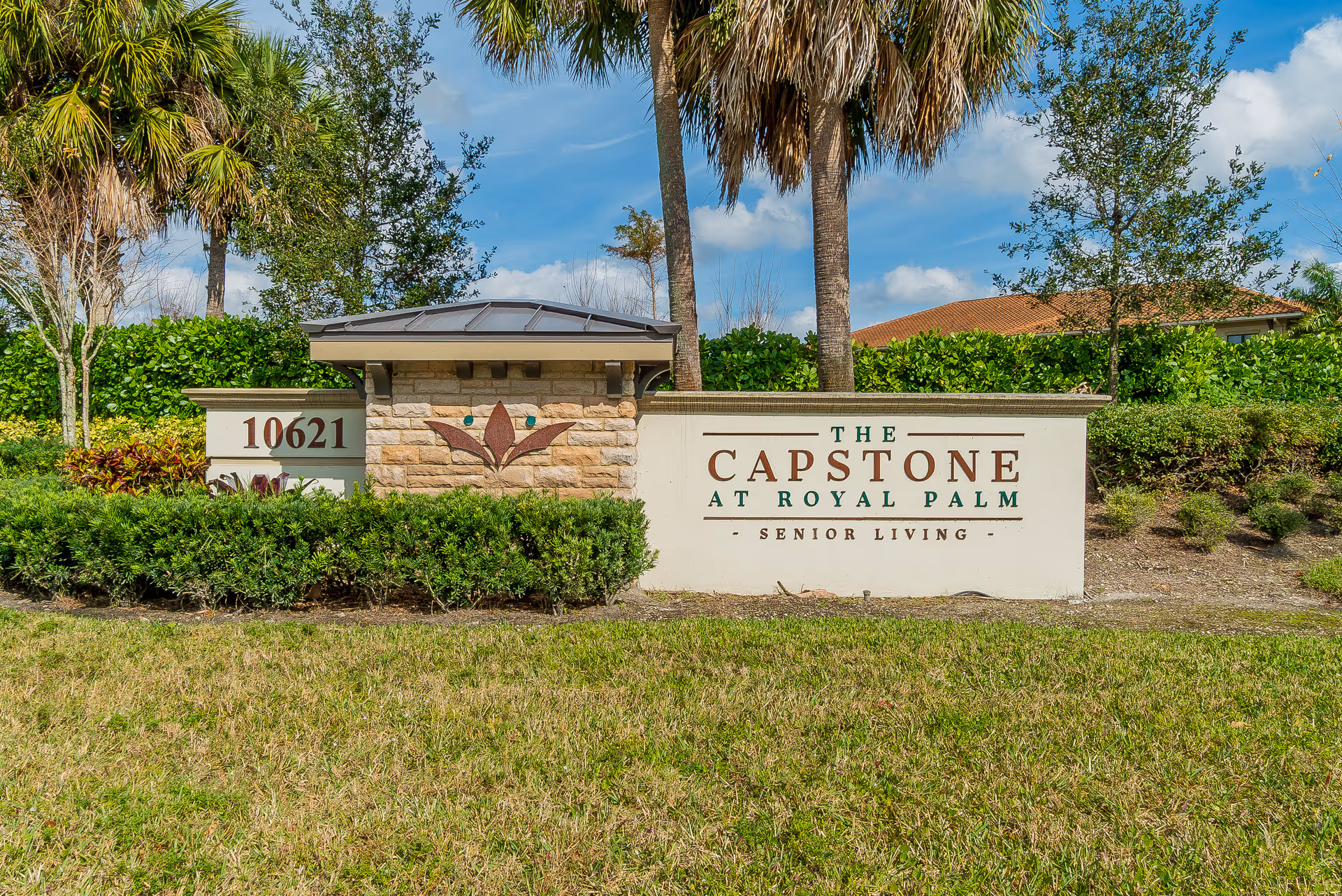 Outdoor view of a landscaped entrance sign for The Capstone at Royal Palm Senior Living, featuring greenery, palm trees, and a clear blue sky.