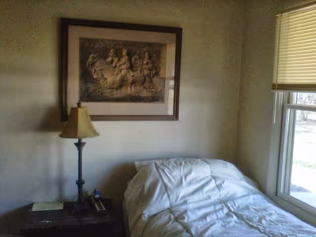 A small bedroom with a single bed covered in white bedding positioned next to a window with blinds. A nightstand beside the bed holds a lamp, a phone, and some small items. Above the nightstand hangs a framed picture depicting a group of people on horseback.