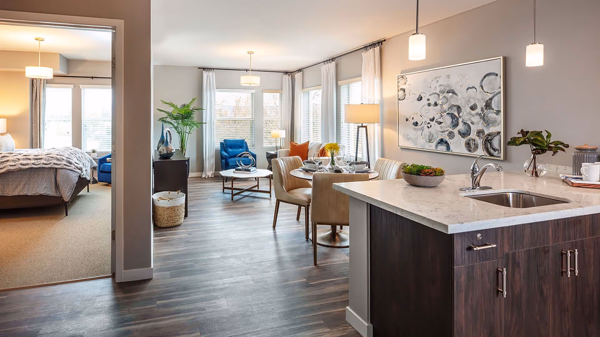 A modern open-concept living space featuring a kitchen island with a sink and pendant lights, a dining area with a round table set for four, and a living room with a blue armchair, a coffee table, and large windows with white curtains. To the left, a bedroom with a bed, knitted blanket, and windows is visible through an open doorway.