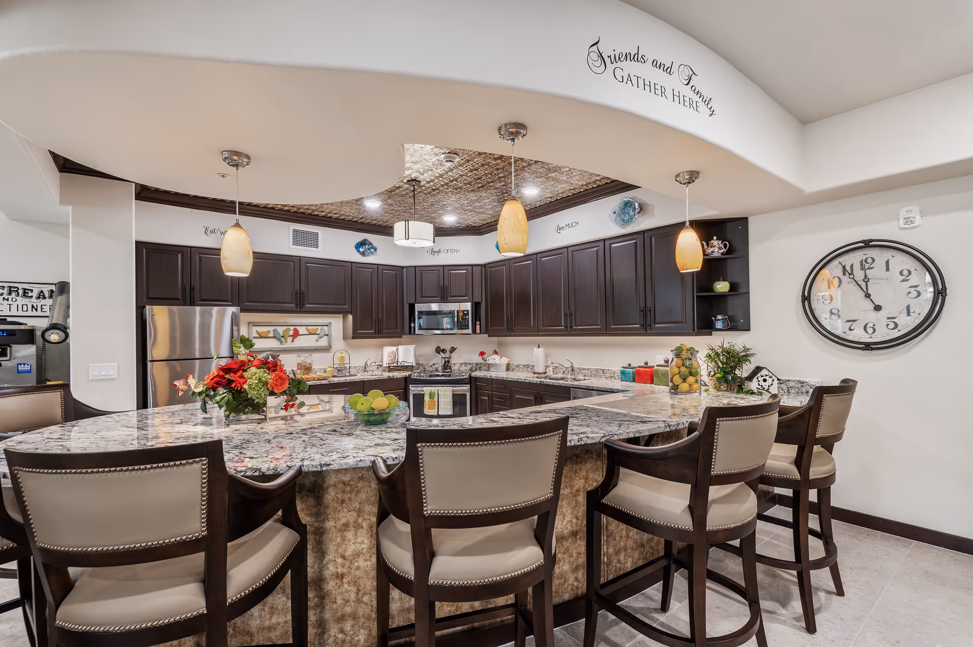 Modern kitchen with dark wood cabinets, granite countertops, and a large island with five cushioned bar stools. The kitchen features stainless steel appliances including a refrigerator and microwave. Pendant lights hang above the island. Decorative items include a bowl of green apples, a vase with flowers, and a large wall clock. Above the cabinets, there is a wall decal that reads 'Friends and Family Gather Here.'