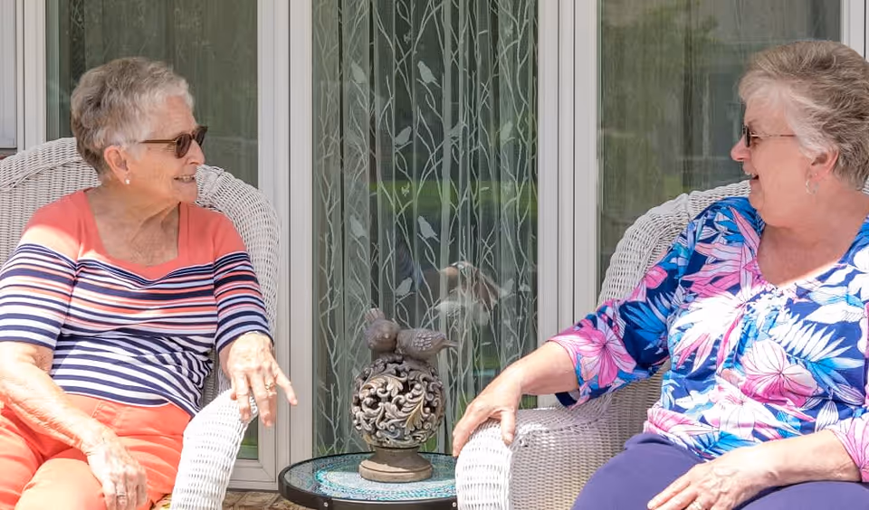 Two elderly women sitting on white wicker chairs on a porch, engaged in conversation and smiling. Between them is a small round table with a decorative bird sculpture. Behind them are large windows with patterned curtains.