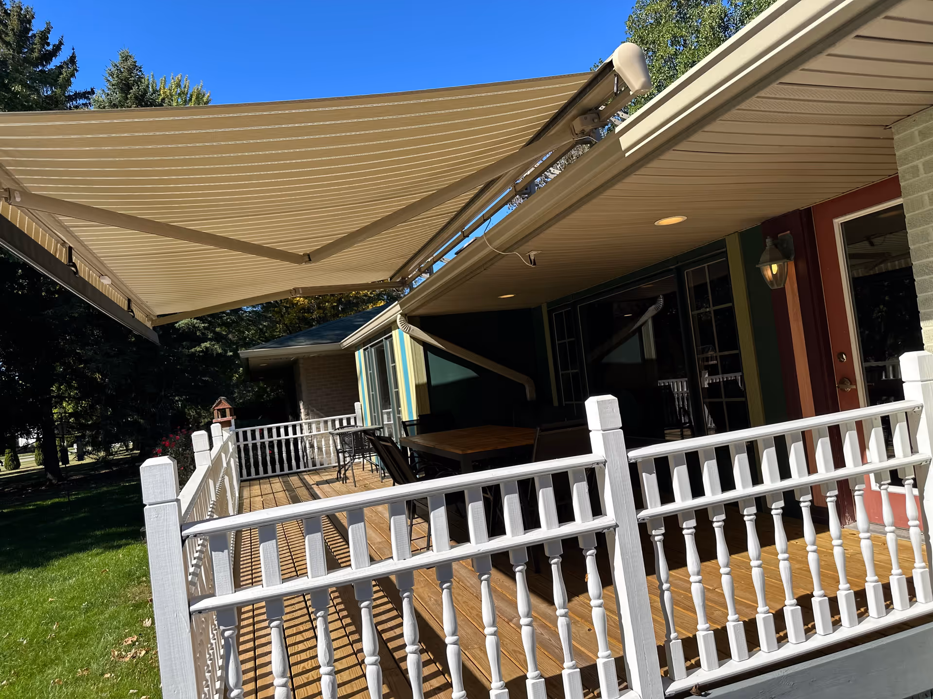 A wooden deck attached to a building with white railing and a retractable beige awning providing shade. There is outdoor furniture including a table and chairs on the deck. The building has a red door and windows, and the area is surrounded by green grass and trees under a clear blue sky.