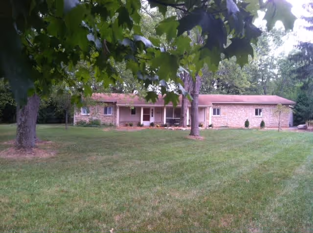 Single-story building with a stone facade and a red roof, surrounded by a large grassy lawn and several trees in the foreground.